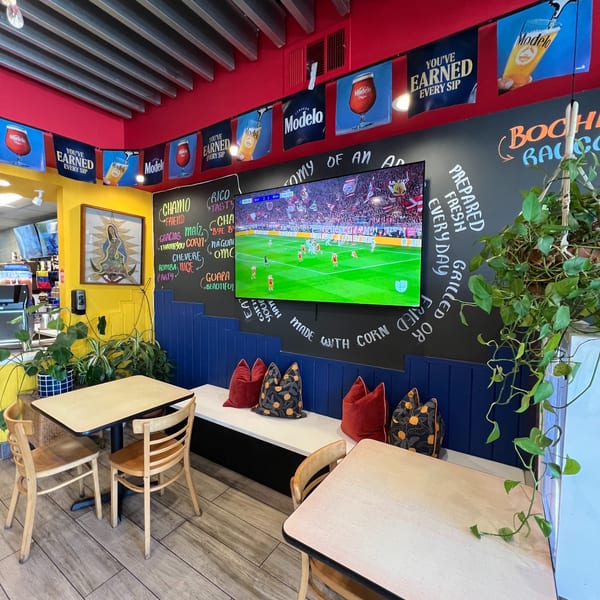 Colorful restaurant interior with a large TV showing soccer, tables, benches, and Modelo beer banners.
